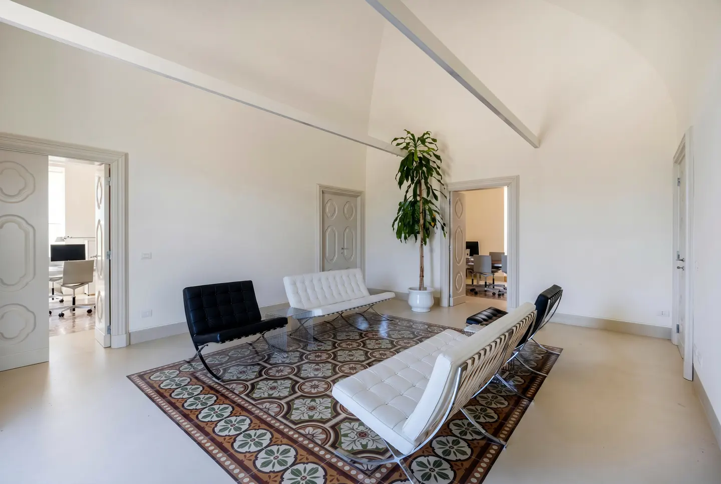 Modern office lounge with white vaulted ceilings, patterned tile rug, two white leather sofas, and two black leather chairs.