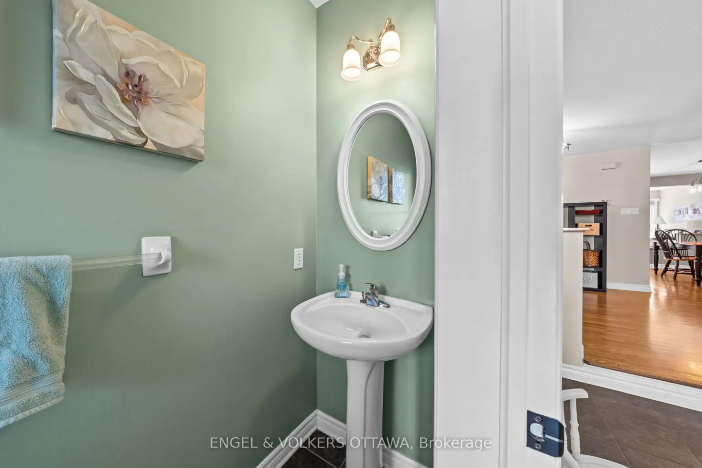 A powder room with green walls, a white pedestal sink, and an oval mirror. A floral painting hangs above a towel bar with a blue towel. A glimpse of the dining room is visible.