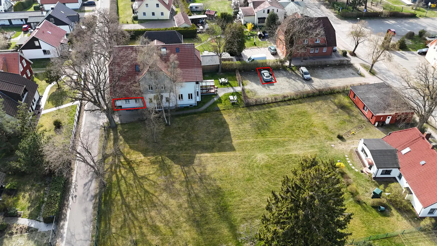 Aerial view of a property with a large lawn, a two-story house with a red roof, and a parking area with cars.