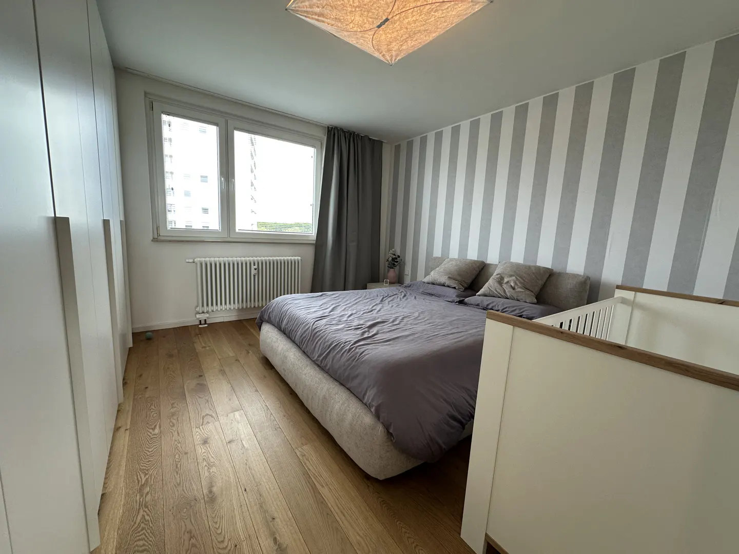 Bedroom with a gray bed, striped wallpaper, and a white crib in the foreground. A window and a radiator are visible.