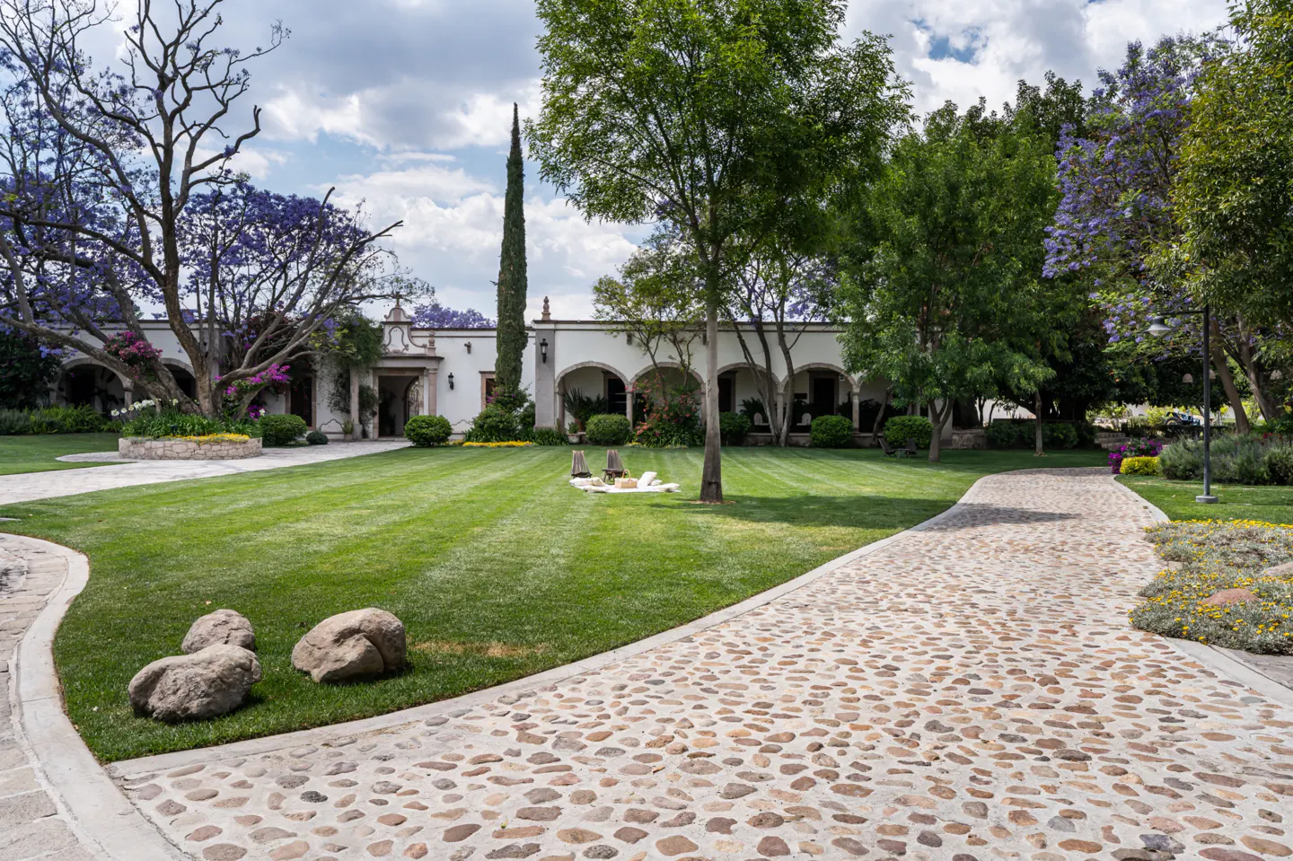 Exterior view of a white hacienda-style home with a stone path, green lawn, trees, and a picnic blanket.