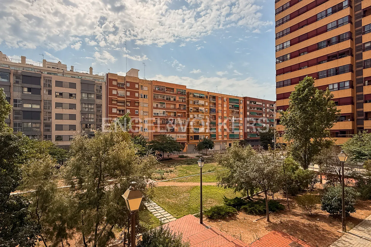 Cityscape view of apartment buildings surrounding a park with trees and lampposts under a cloudy sky.