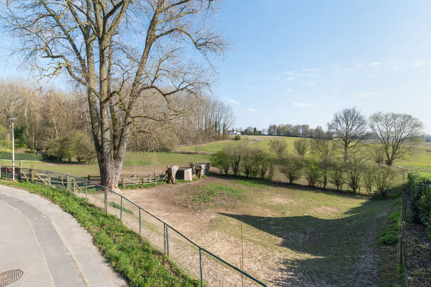 A landscape view of a field with trees, bushes, and a fence under a blue sky.