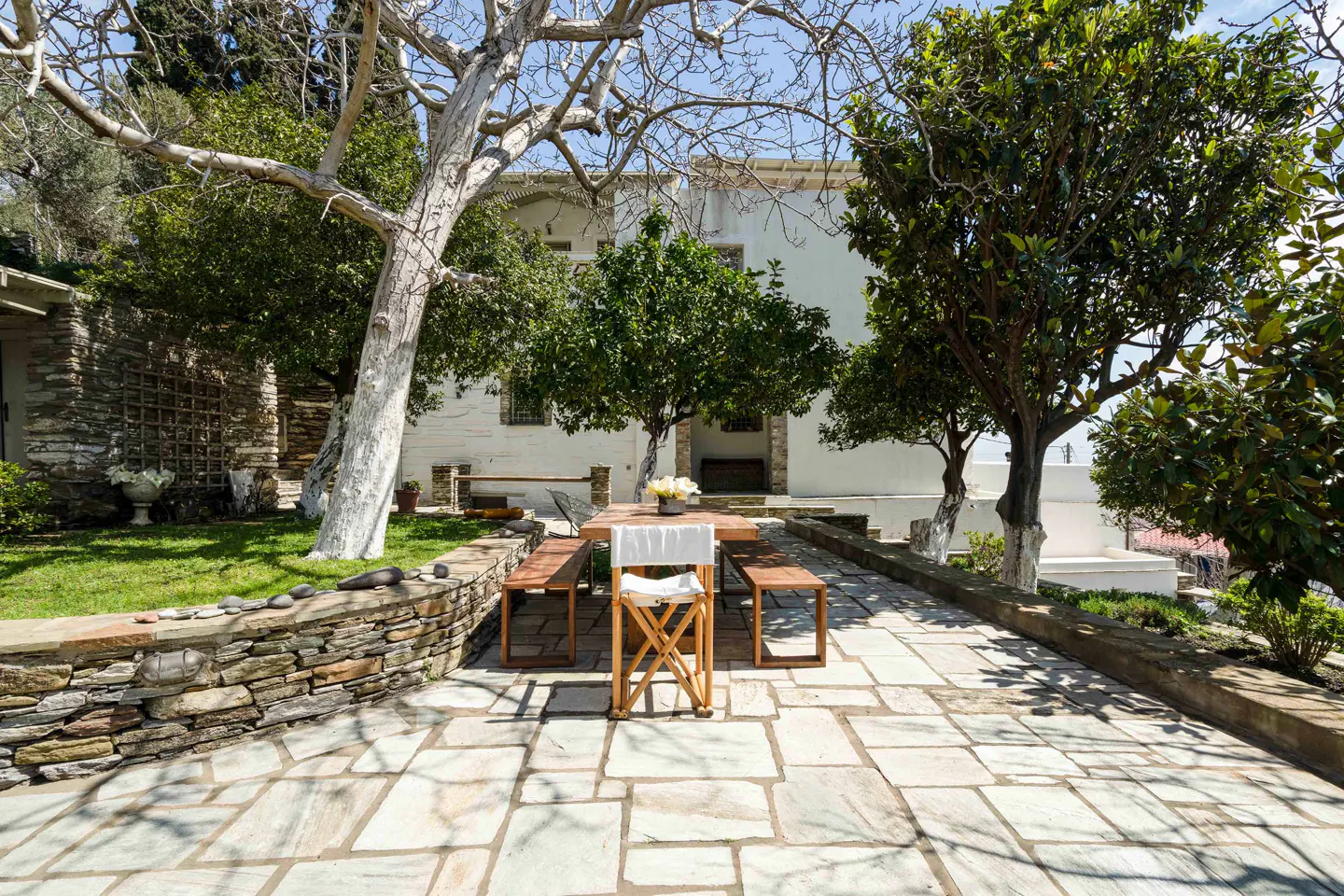 Outdoor patio with stone tiles, a wooden table, benches, and a white director's chair. Green trees surround the area, with a white building in the background.
