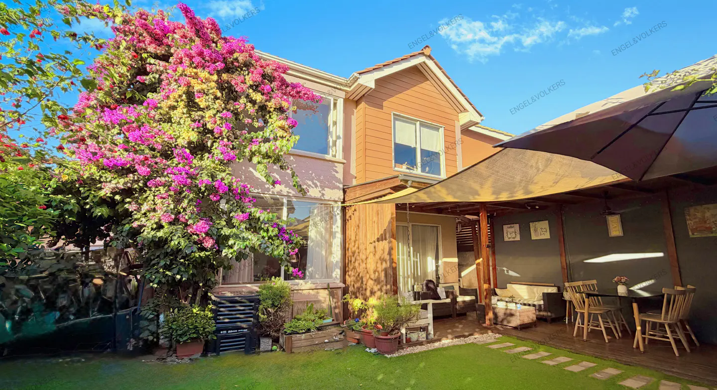 Two-story house with pink bougainvillea, a patio with a table, chairs, and a sunshade.