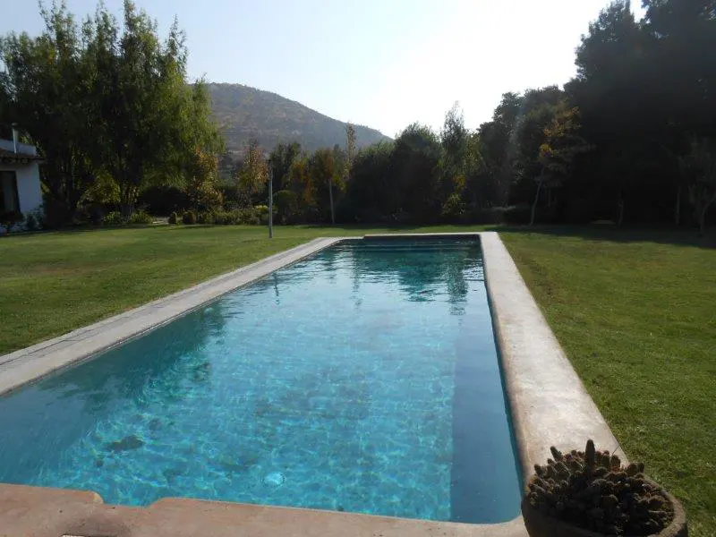 A rectangular swimming pool with turquoise water, surrounded by green grass and trees. A mountain is visible in the background.