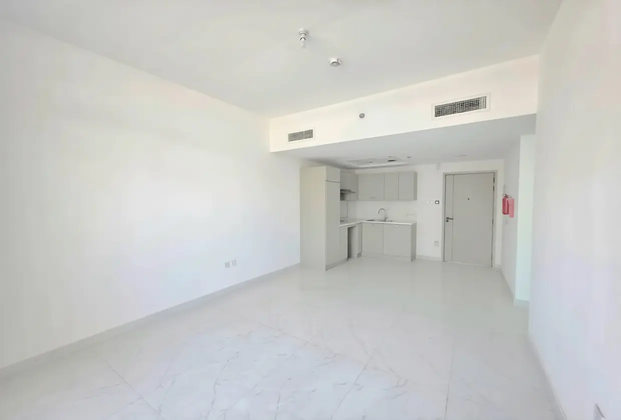 Bright, empty apartment with white walls, marble floors, and a small gray kitchen area. A gray door is visible in the background.