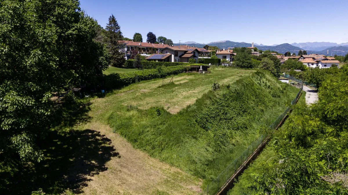 A grassy lot with a fence, trees, and houses with red roofs in the background on a sunny day.