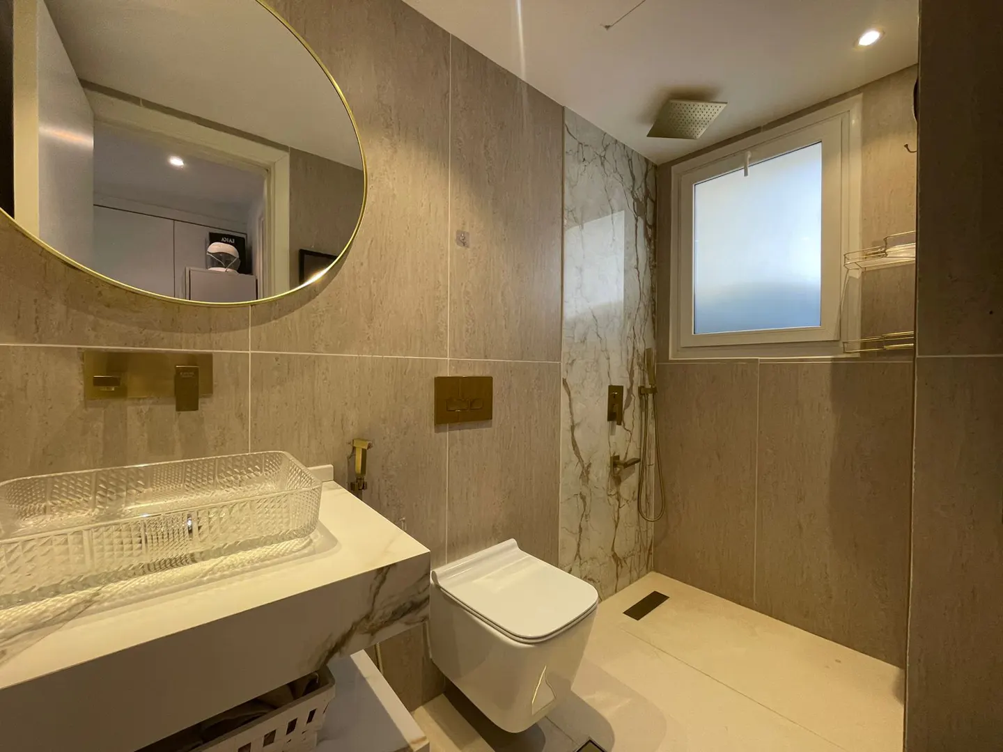 Modern bathroom with beige tile walls, white toilet, marble sink, and gold fixtures. A round mirror hangs above the sink.