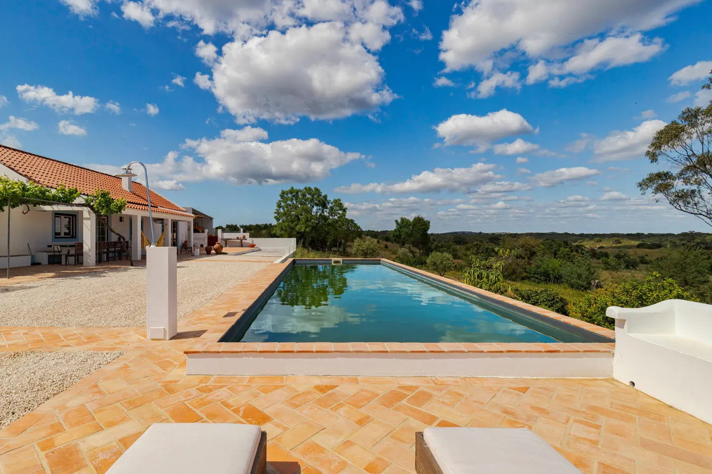 Outdoor pool with blue water, surrounded by orange brick patio. White house with orange roof in background under a blue sky with white clouds.