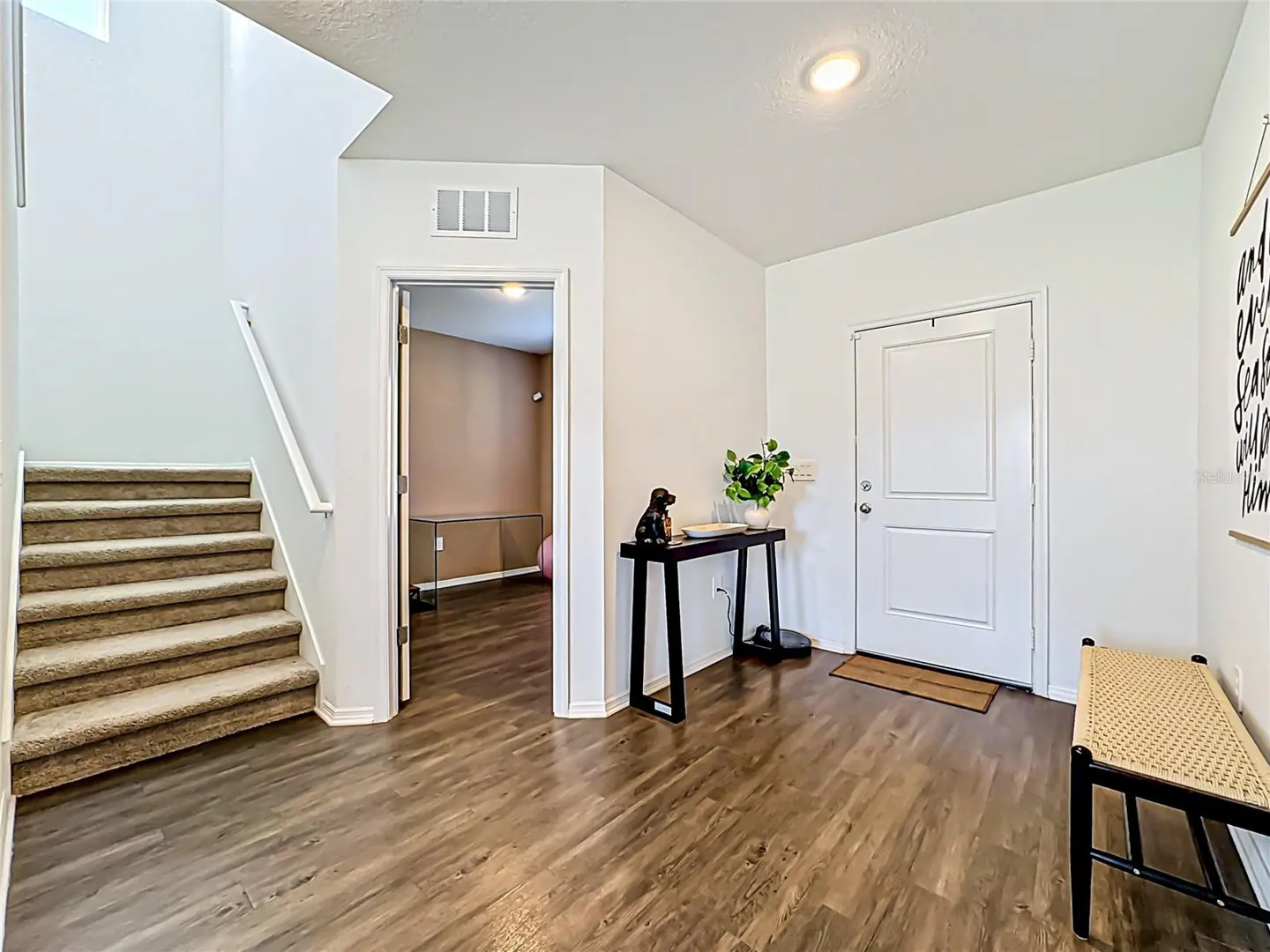 Bright entryway with wood-look floors, white walls, and a carpeted staircase. A black console table with a plant sits near the white front door. A woven bench is on the right.