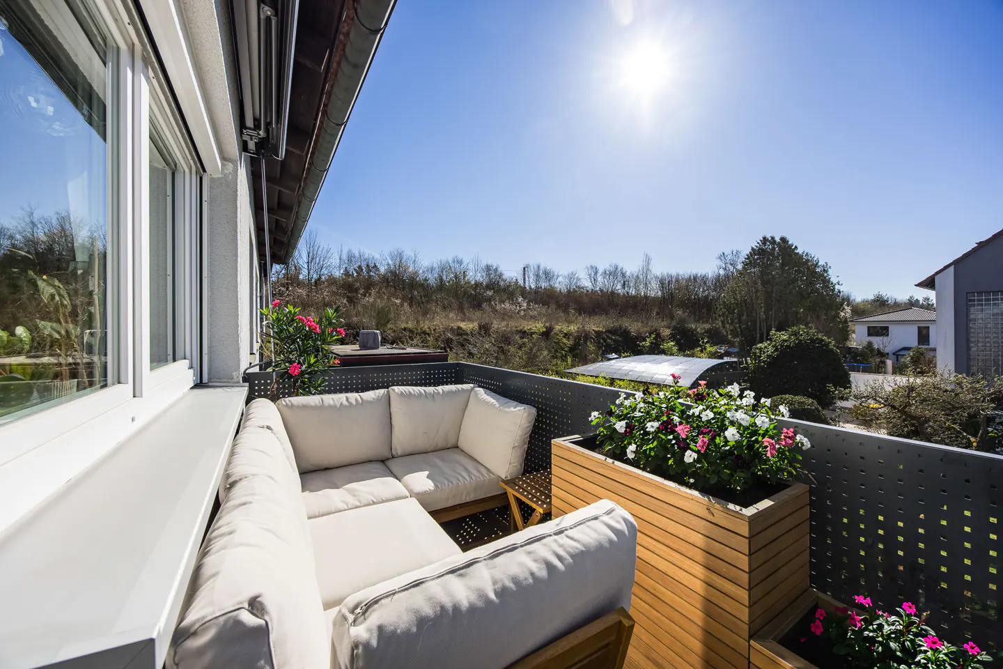 Sunny balcony with a white cushioned sofa, wooden planters with flowers, and a view of trees and a blue sky.