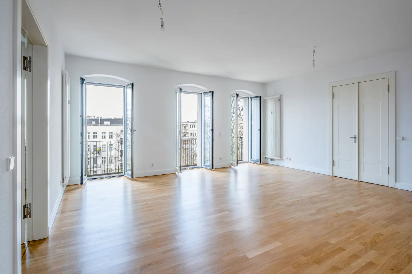 Bright, empty room with hardwood floors, white walls, and three open balcony doors showing city views.