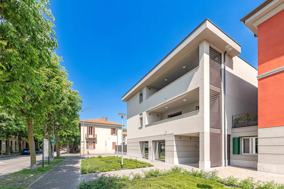 Modern two-story apartment building with balconies, next to a sidewalk and green trees under a clear blue sky.