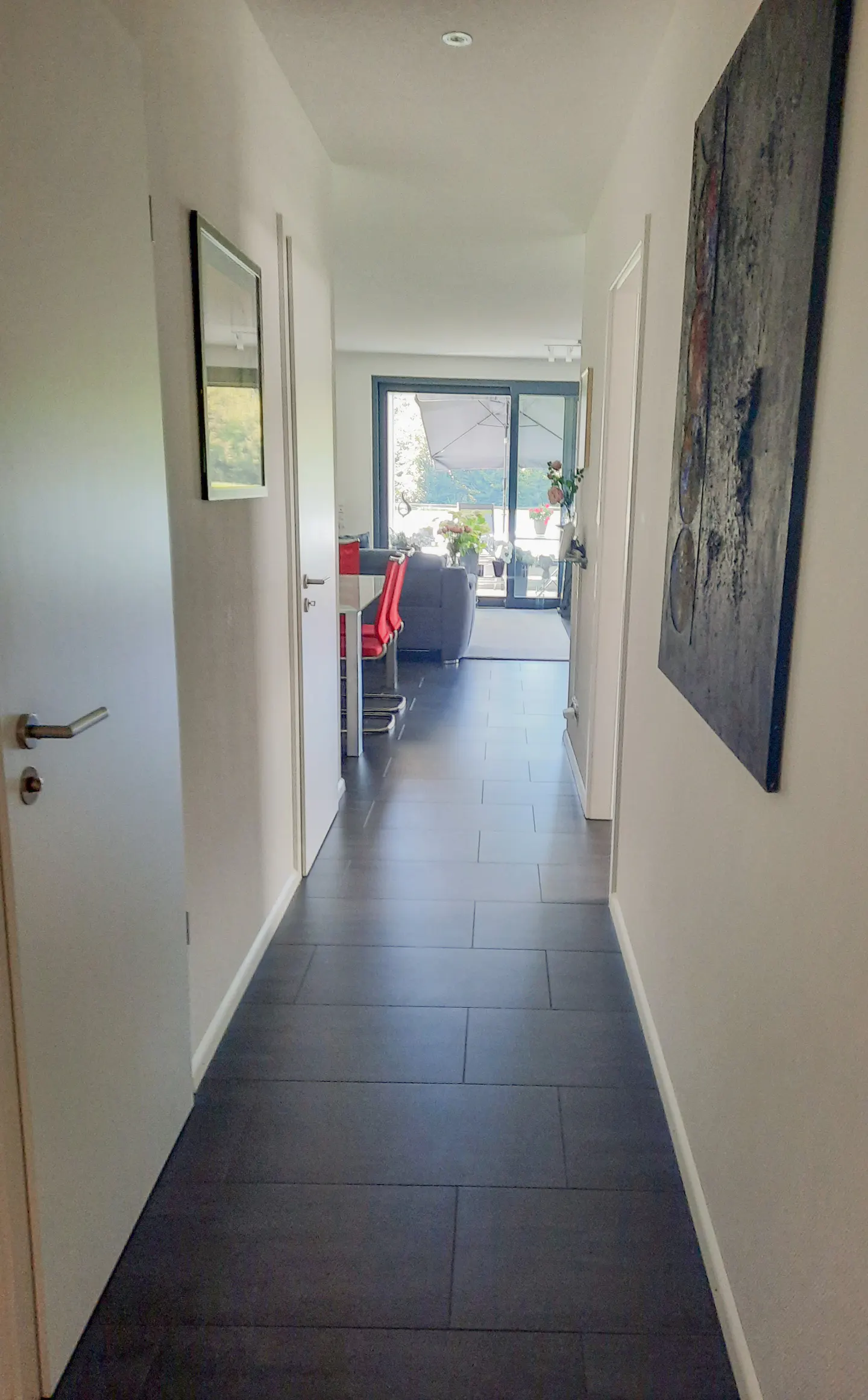 Hallway view with dark tile floor, white walls, and doors. Living room visible with gray sofa and red chairs. Abstract art on the right wall.
