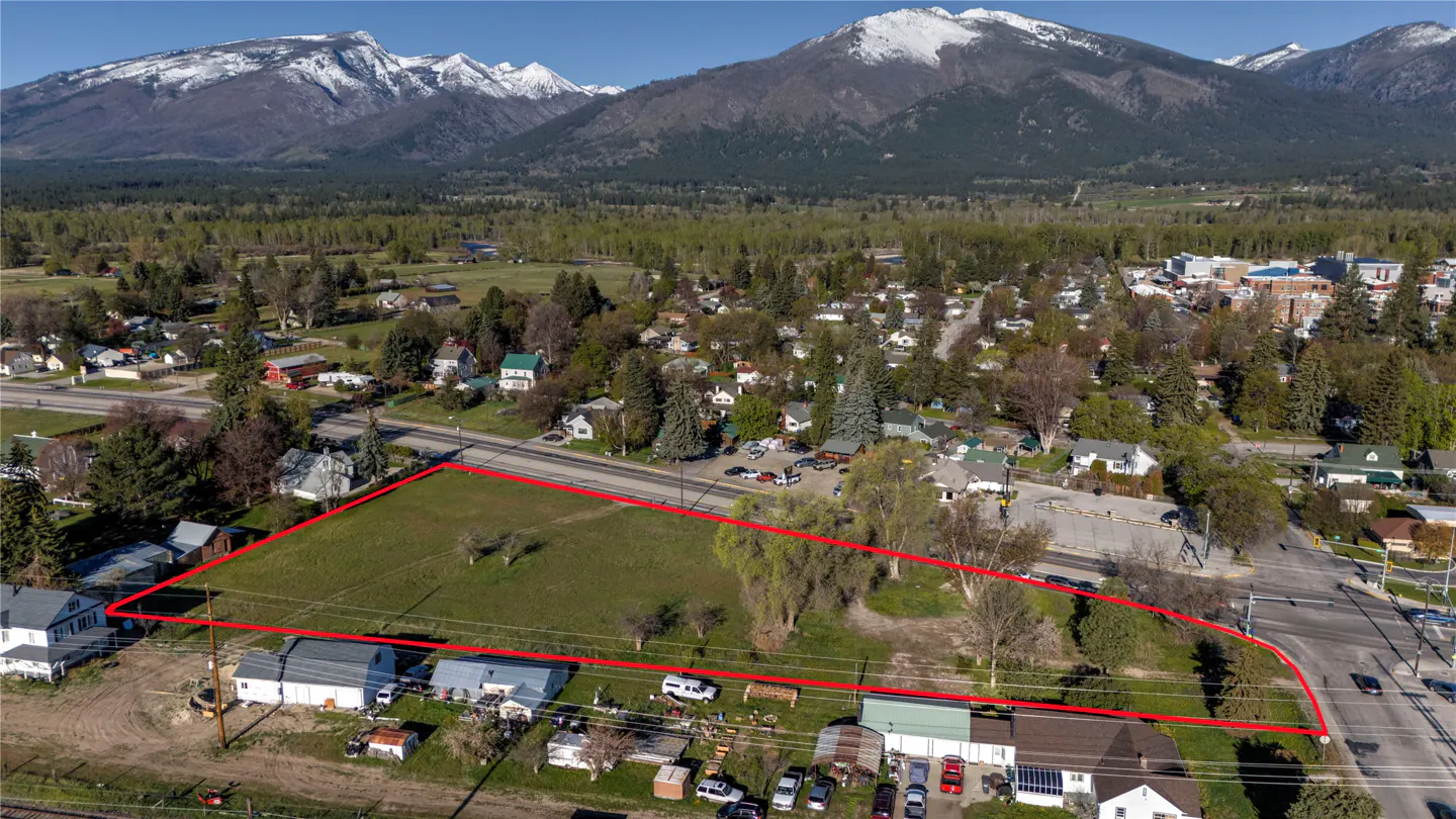 Aerial view of a grassy lot outlined in red, with houses, trees, and snow-capped mountains in the background.