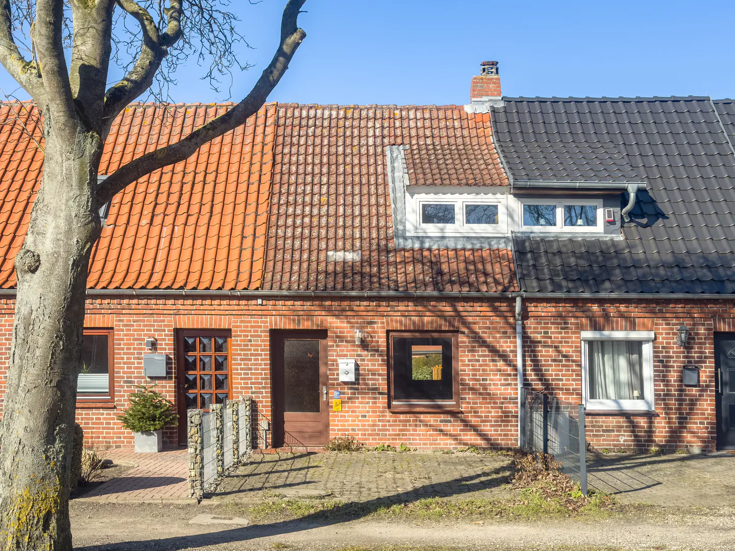 Two brick townhouses with red and black tiled roofs under a blue sky, separated by a tree.