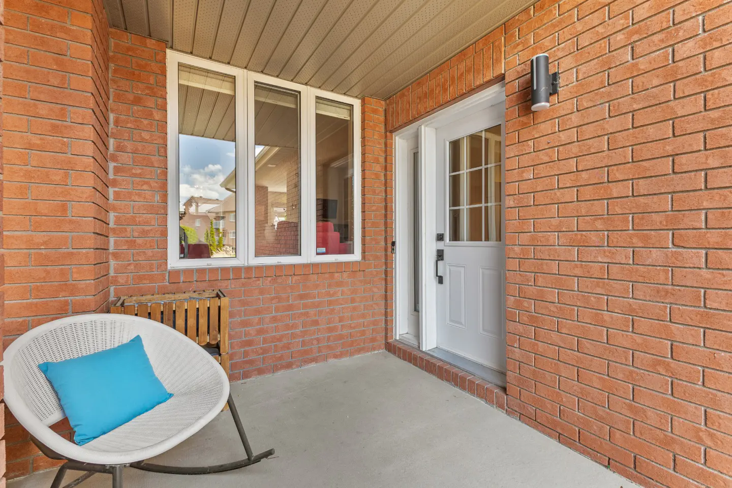 A white rocking chair with a blue pillow sits on a porch with red brick walls, a white front door, and large windows.