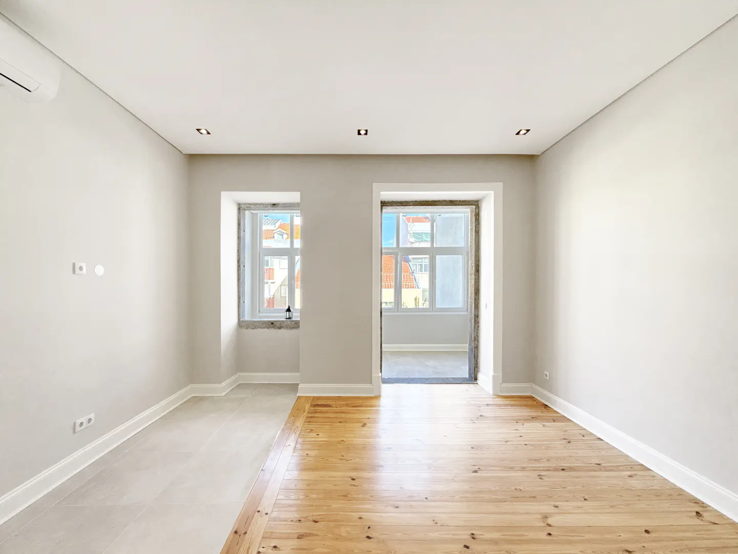 Bright, empty room with light gray walls, wood floors, and white trim. A window and doorway show a view of buildings outside.