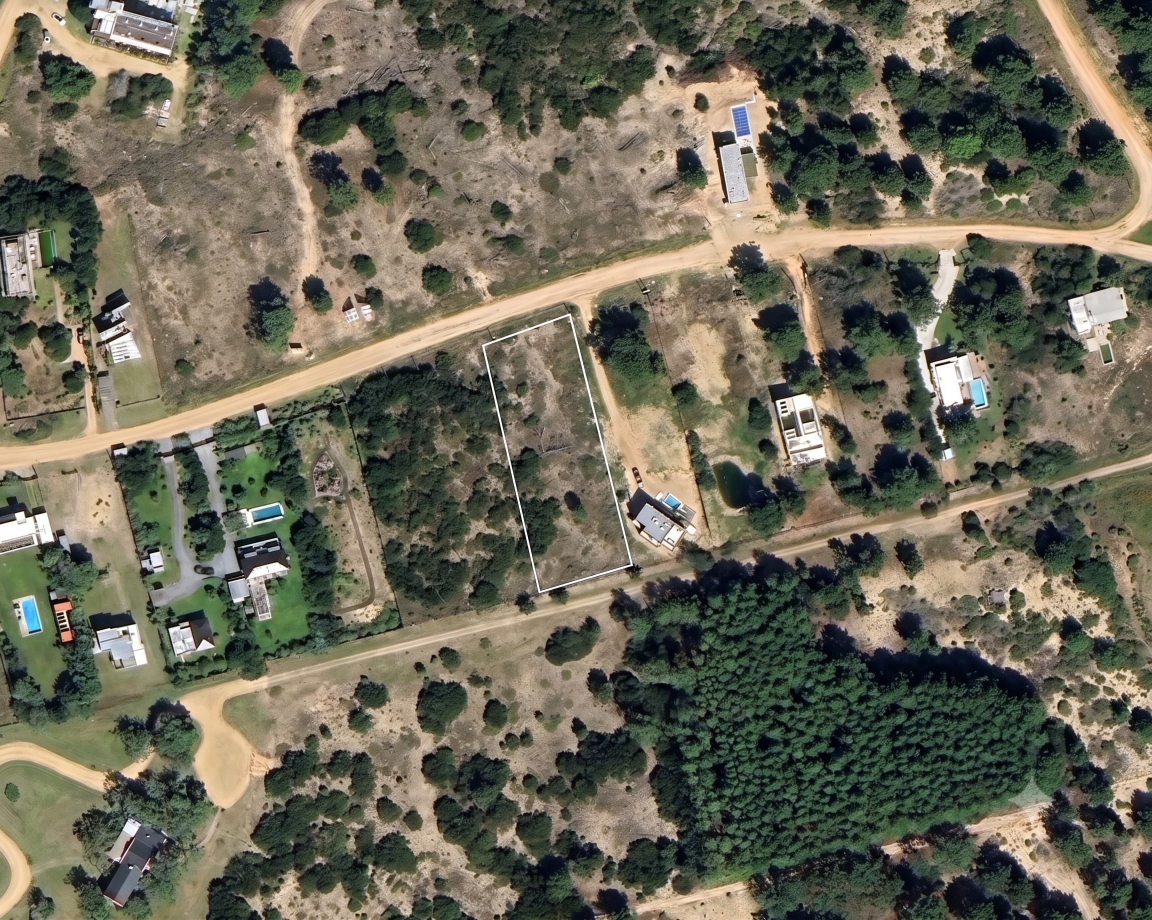 Aerial view of a rectangular vacant lot outlined in white, surrounded by trees, dirt roads, and nearby residential homes.