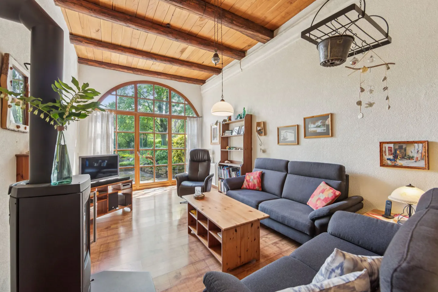 Living room with wood floors, exposed beams, and a large arched window overlooking a green garden. Gray sofas and a wood coffee table furnish the space.
