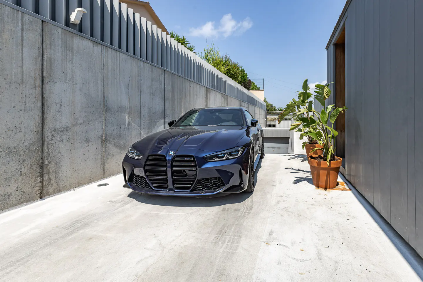 A dark blue BMW parked on a concrete driveway between a tall concrete wall and a modern building with potted plants.