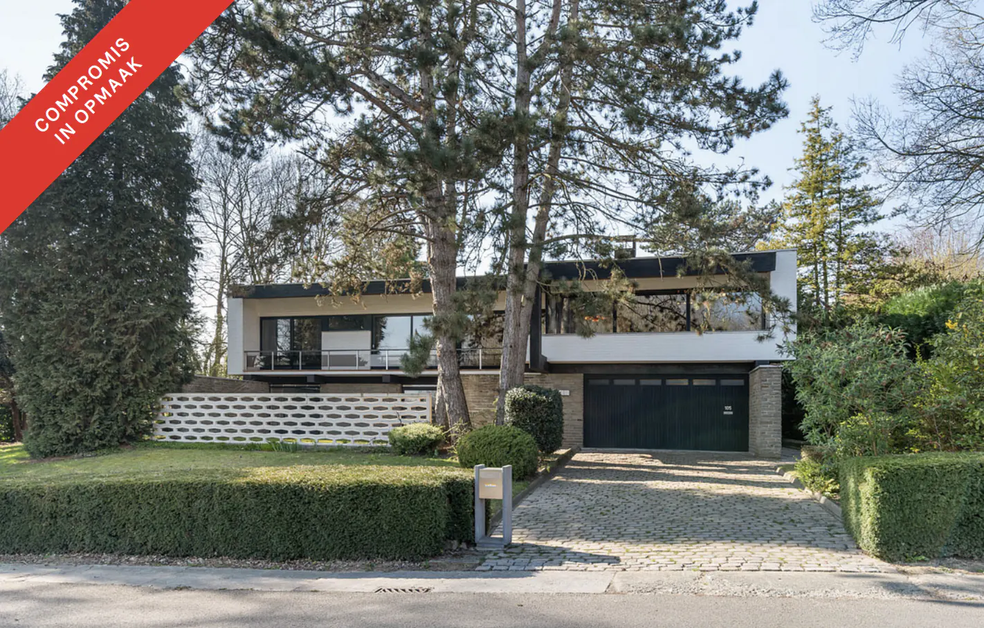Two-story modern house with a black garage door and large windows, surrounded by trees and a green lawn. A red banner is in the upper left corner.