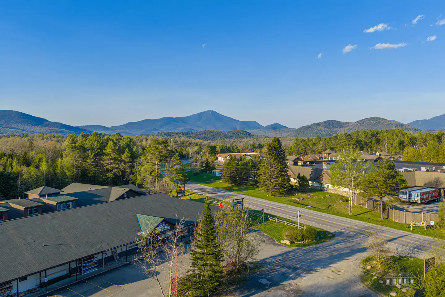 Aerial view of a town with a road, buildings, and mountains in the background under a blue sky.