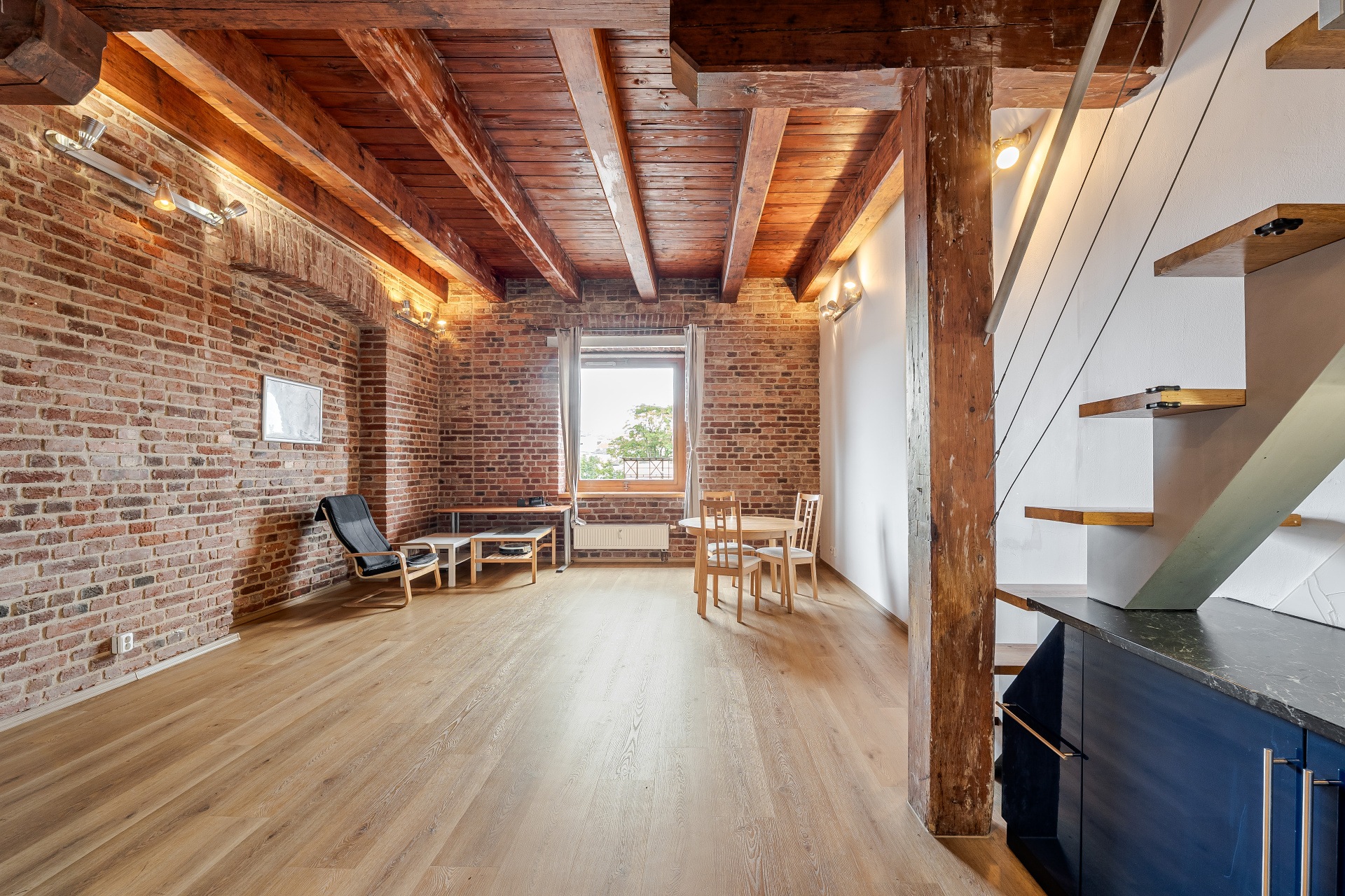 Loft living area with exposed brick walls, wooden ceiling beams, light wood floors, a dining set, and a floating staircase. [AI-generated text]