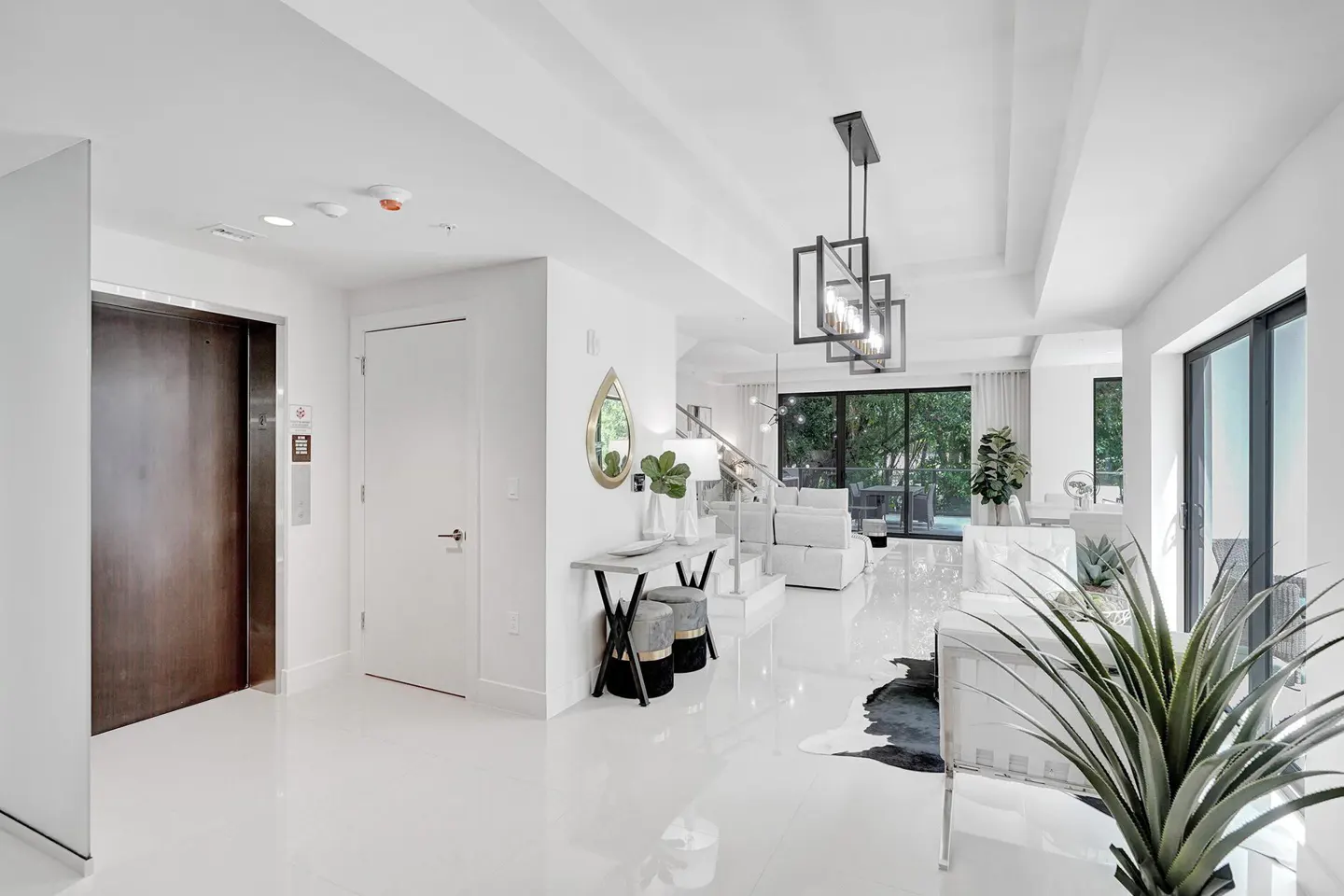 Bright, modern foyer with white walls and floors. An elevator and white door are to the left. A chandelier hangs above the living area.