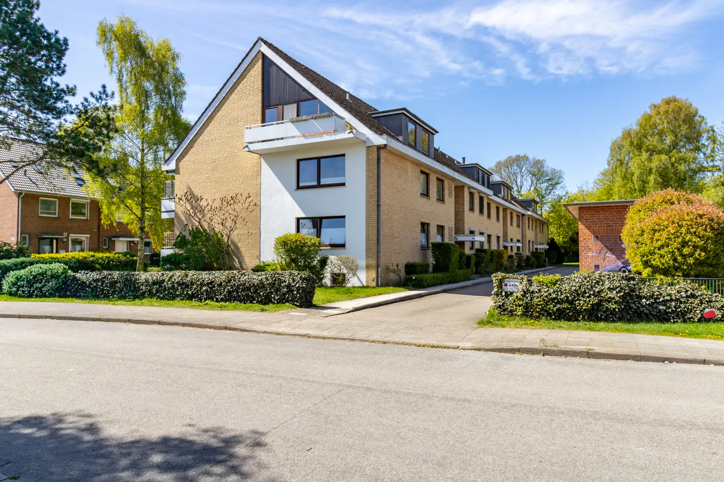 Exterior view of a row of yellow brick townhouses with dark brown roofs and green hedges under a blue sky.