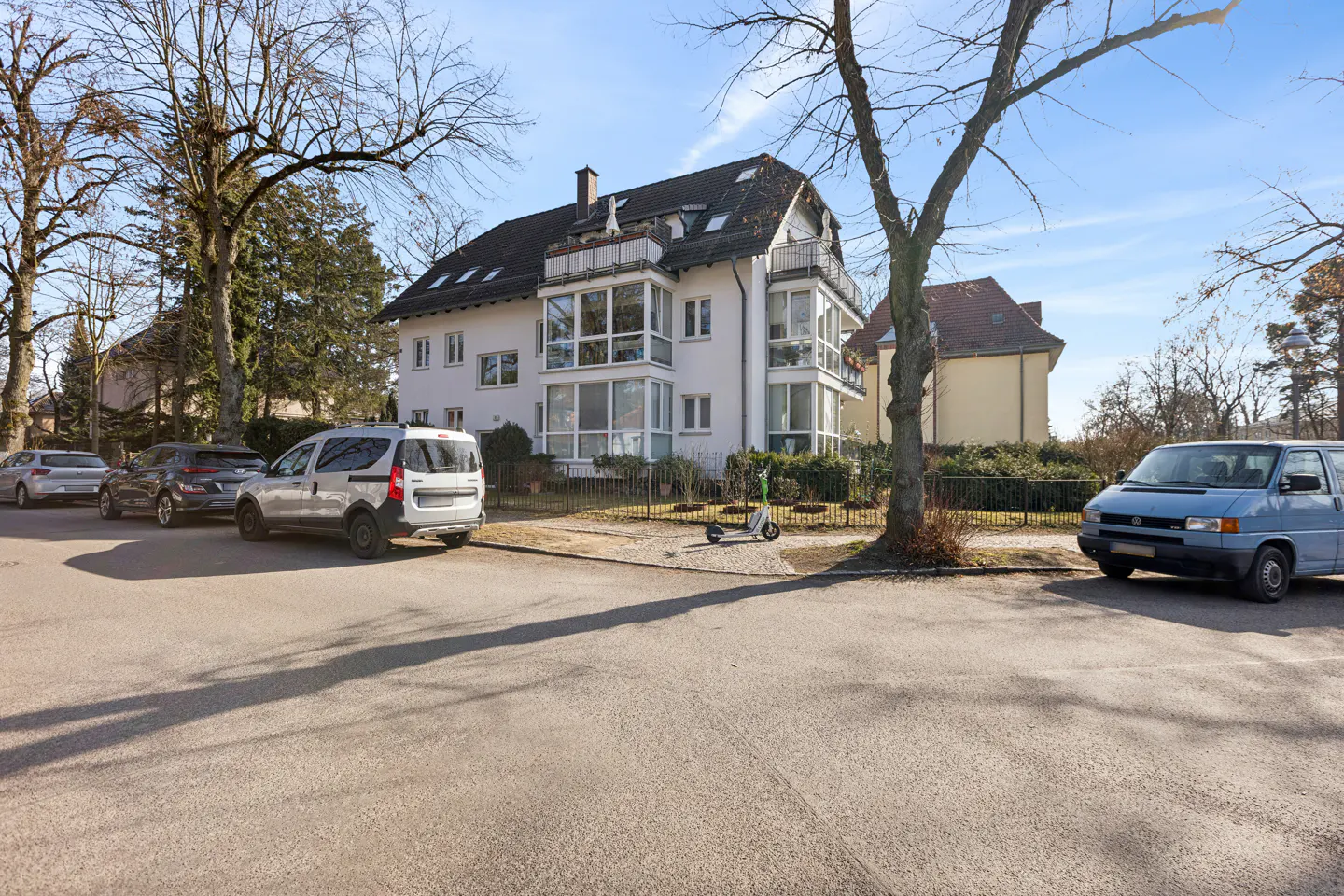 Exterior view of a three-story white house with a black roof, surrounded by trees and parked cars on a sunny day.