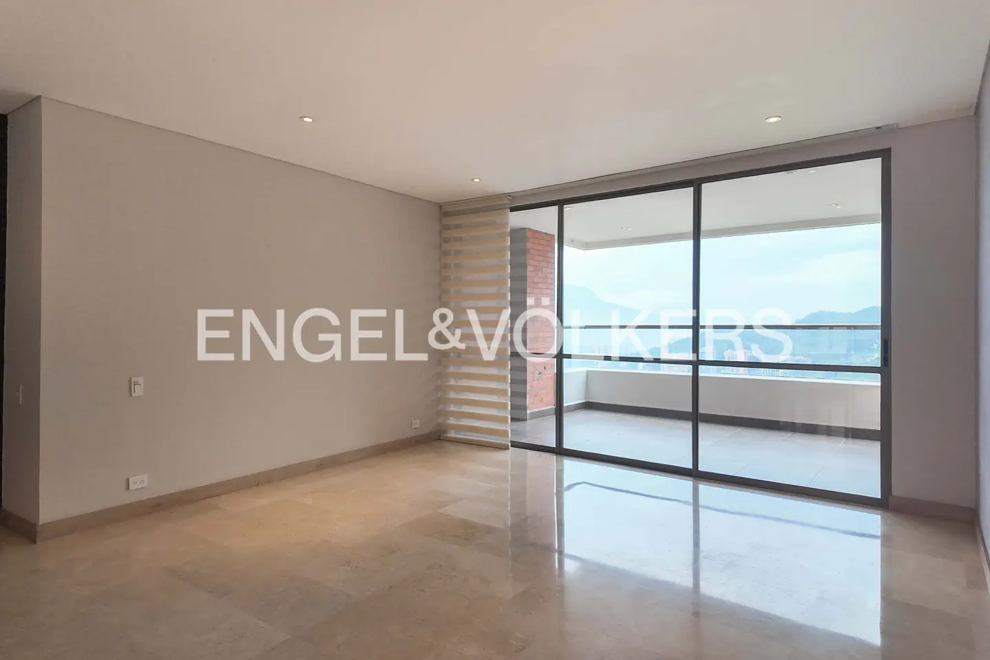Empty room with beige walls, marble floor, and large sliding glass doors to a balcony with a mountain view. Engel & Volkers logo on the wall.