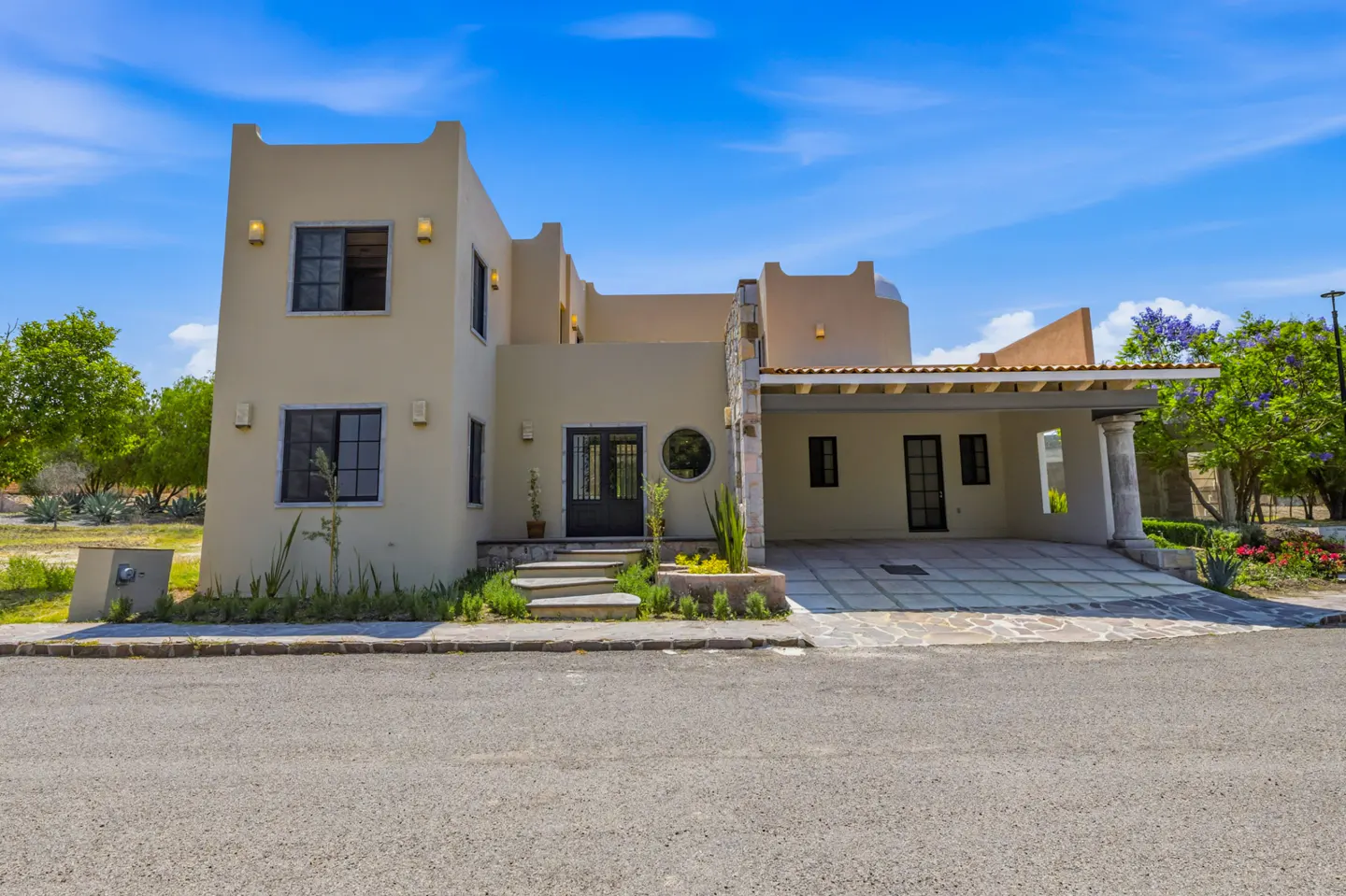 Beige two-story house with a carport, stone accents, and a blue sky background. The house has black framed windows and a black front door.