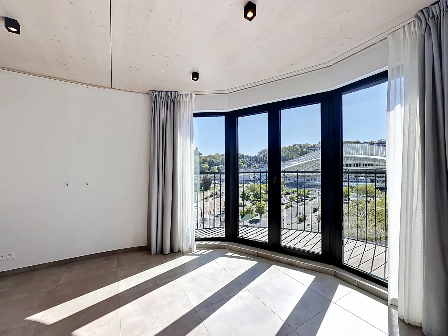 Bright, empty room with tile floor and large black-framed windows. Gray curtains frame the windows, offering a city view with a stadium.