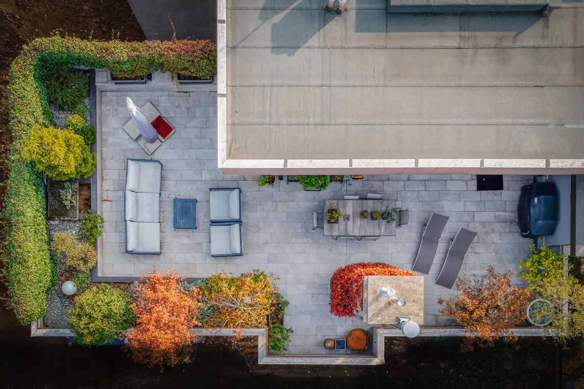 Aerial view of a patio with outdoor furniture, a grill, and lush landscaping. The patio is paved with light gray stone.