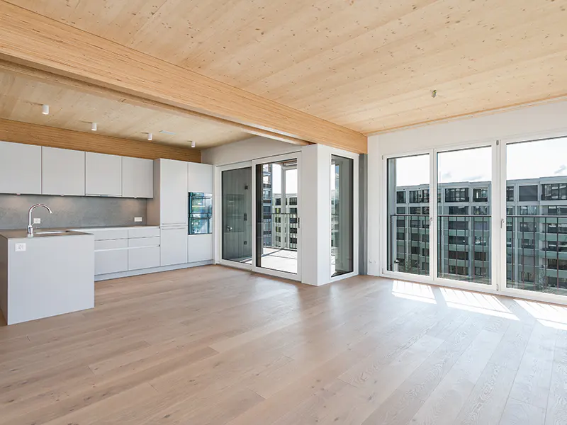 Bright, modern apartment interior with light wood floors, exposed wood ceiling beams, and white kitchen cabinets. Large windows offer city views.