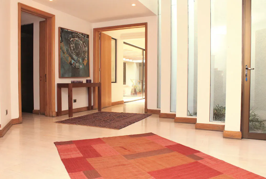 A bright foyer with marble floors, a red geometric rug, and a brown rug. A wooden table with art above it sits near an open doorway. Tall windows line the right wall.