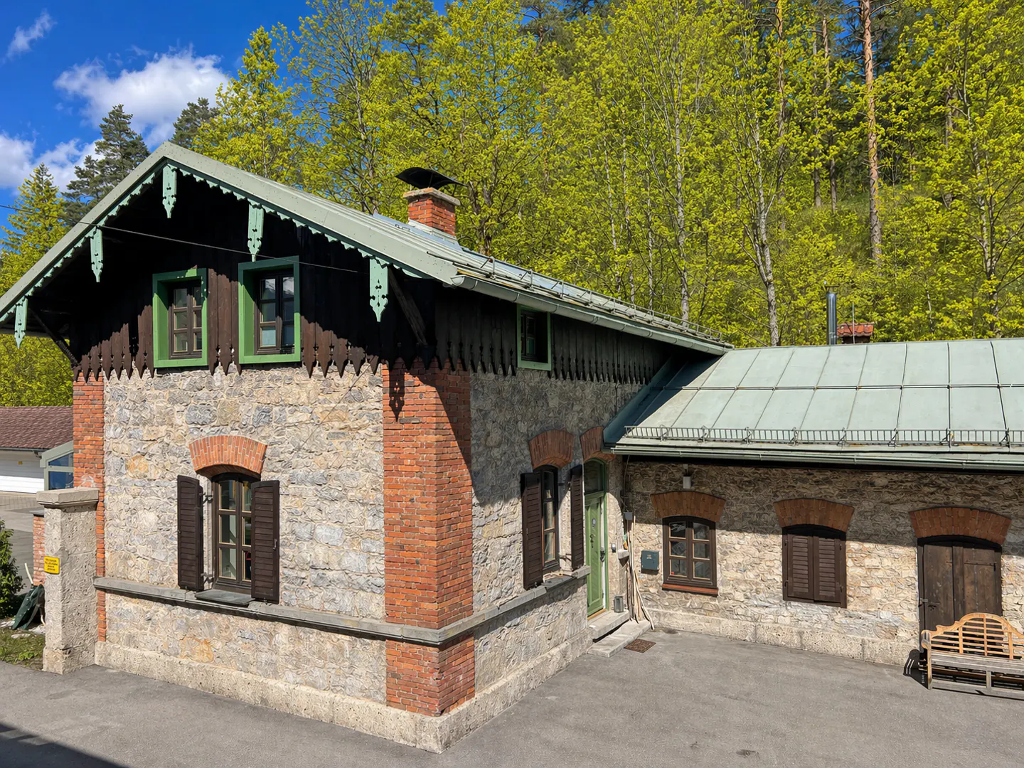 Stone house with brick accents, green trim, and a metal roof, set against a backdrop of lush green trees.