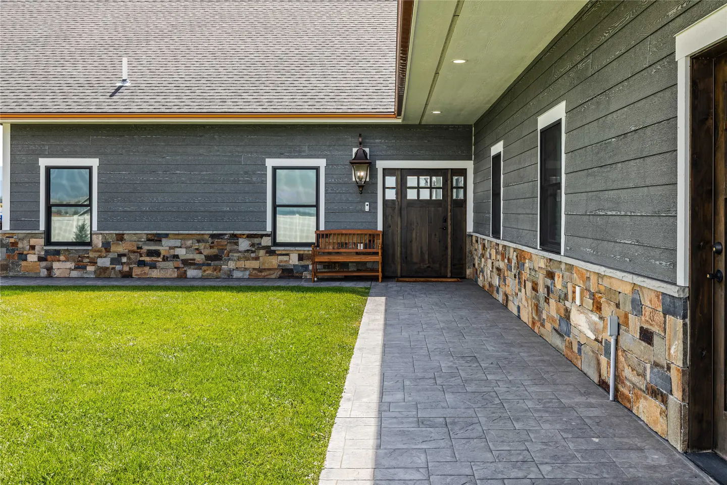 Modern home exterior with gray horizontal siding, stone base, paved walkway, wooden front door, and a bench on the porch.