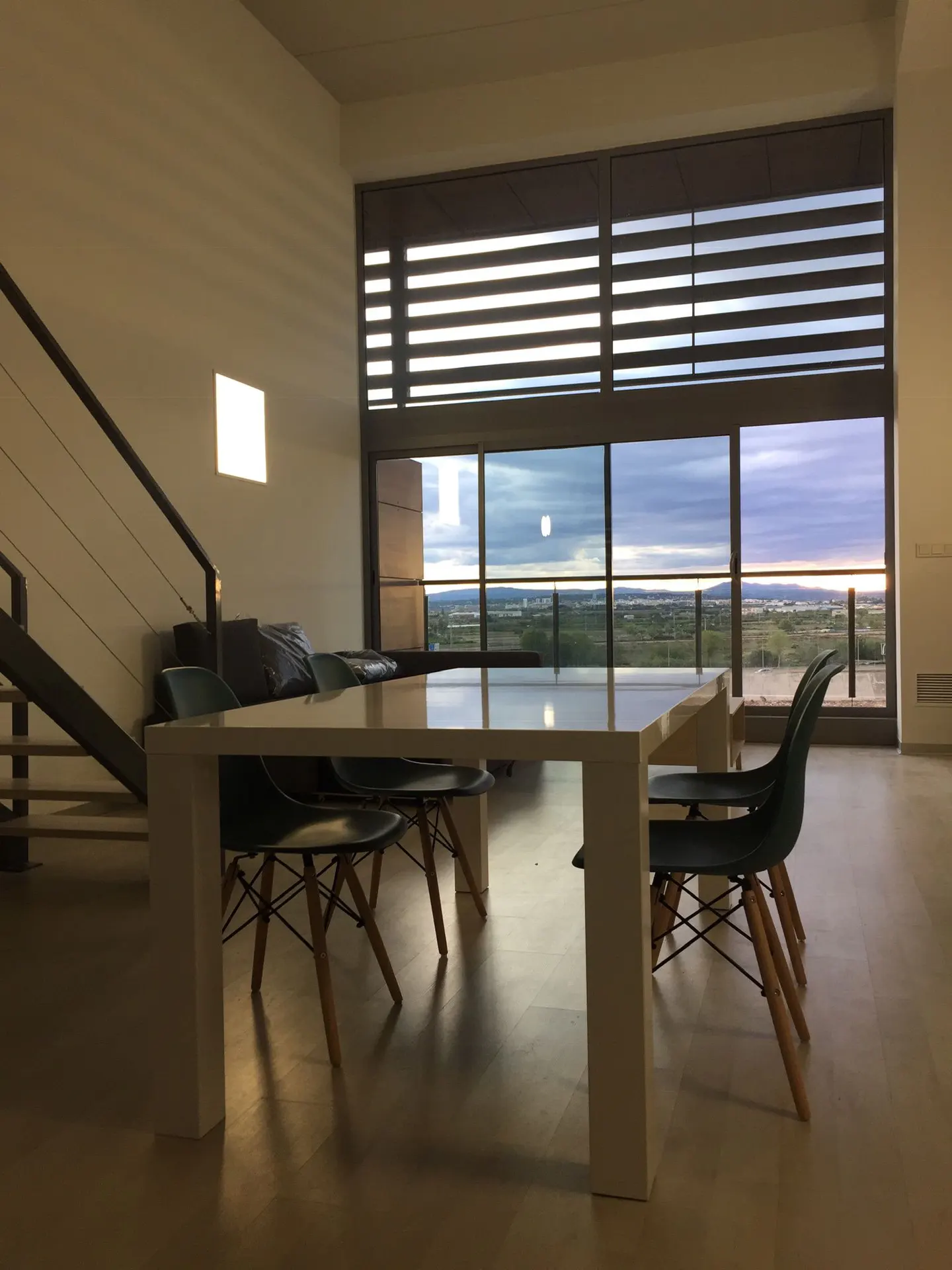 Modern dining area with a white table, dark chairs, and a large window overlooking a scenic landscape at dusk.