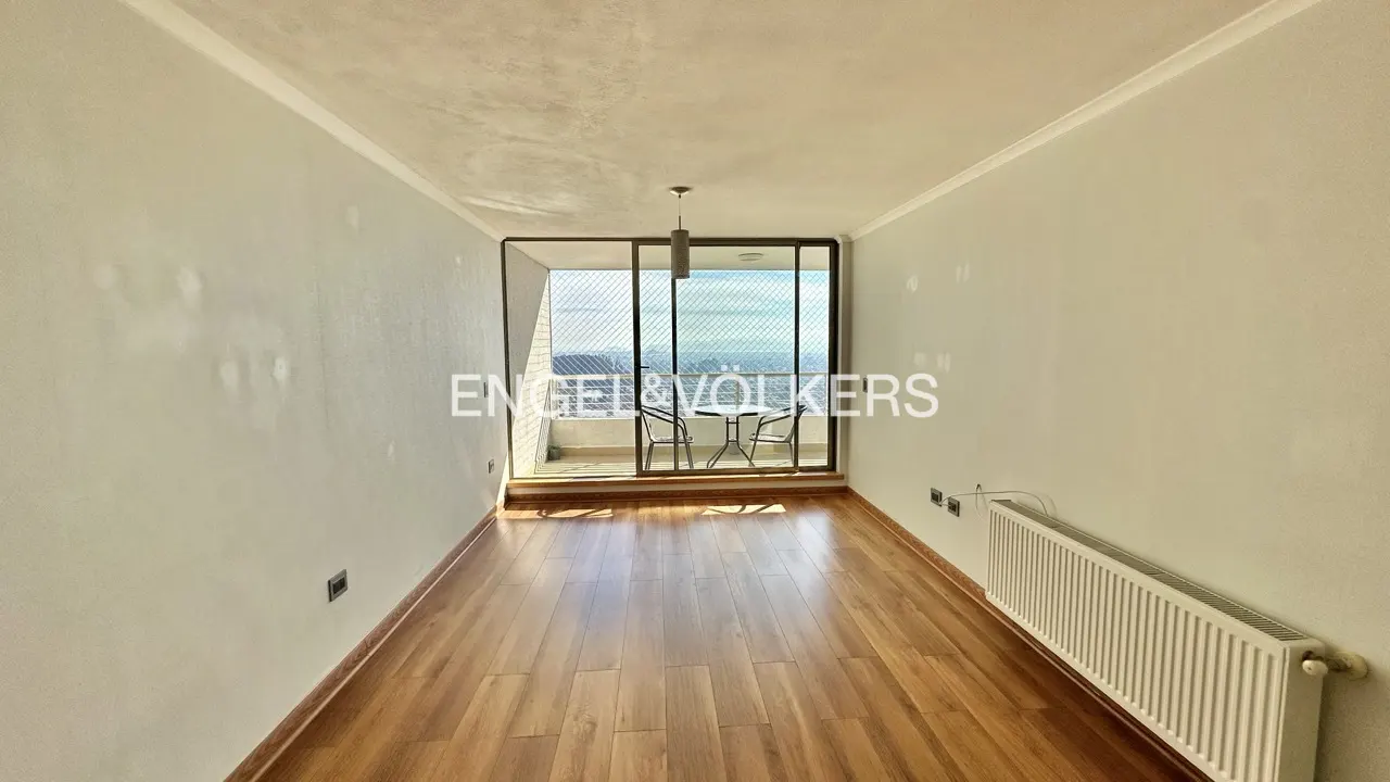 Empty room with wood floors, white walls, and a balcony with a table and chairs overlooking a city view.