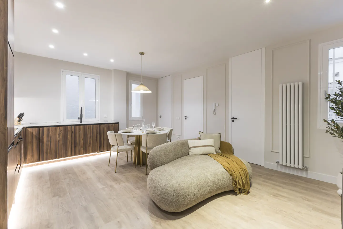 Modern open-plan living area with a curved beige sofa, dining table, and kitchen with wood cabinets and light wood floors.
