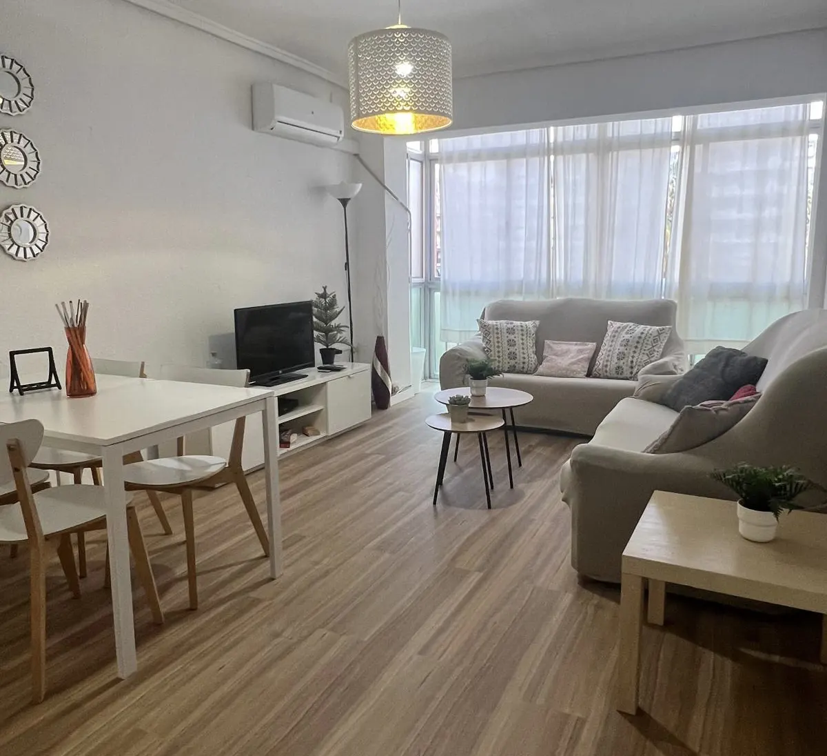 Bright living room with wood floors, white dining set, beige sofa, TV console, and large windows with sheer curtains.