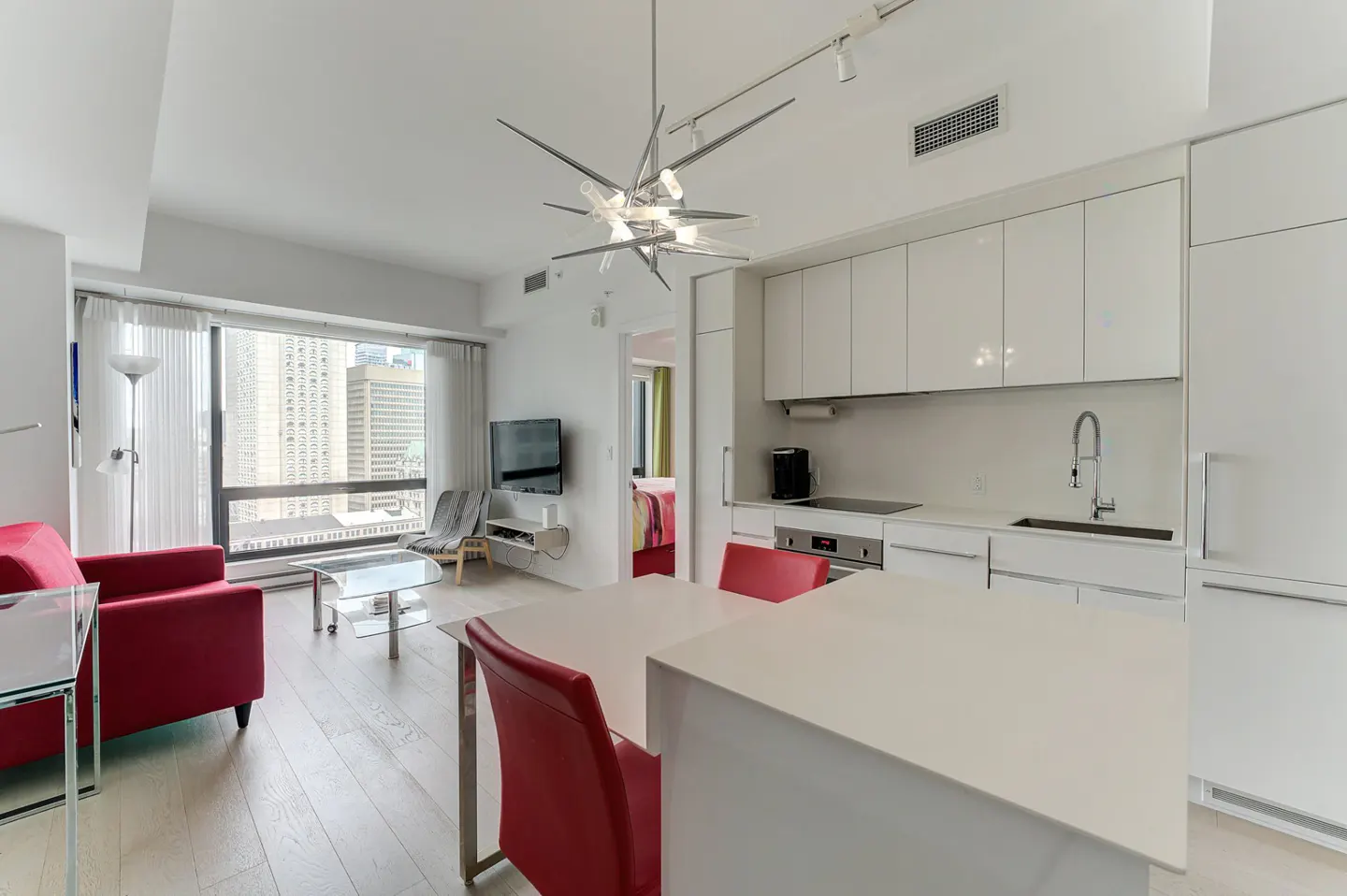 Bright, modern condo interior with white kitchen cabinets, red chairs, and a red sofa. A large window shows a city view.