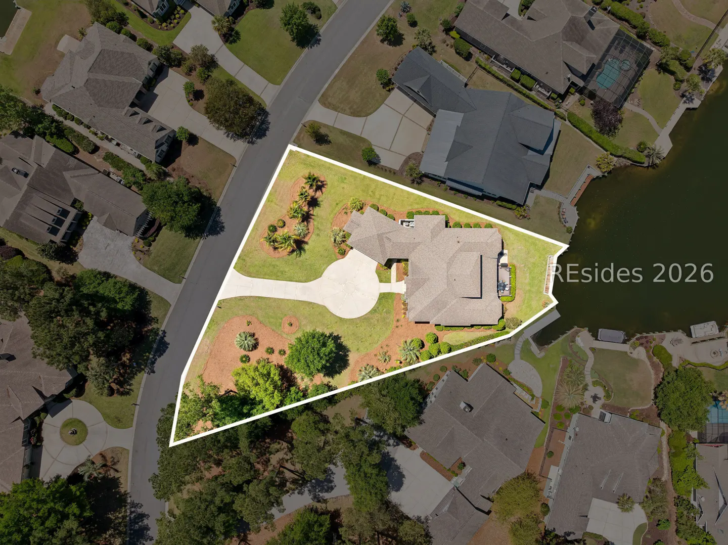 Aerial view of a tan house with a circular driveway, surrounded by green grass and trees, outlined in white.