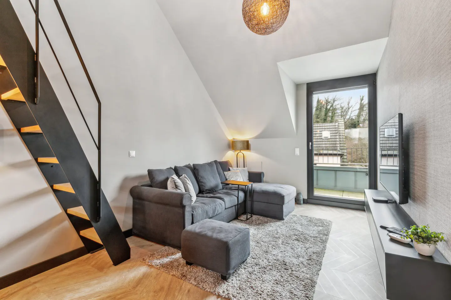 Living room with gray sofa, ottoman, and rug. Black metal stairs, TV, and balcony door. Light wood floors and a woven pendant light.