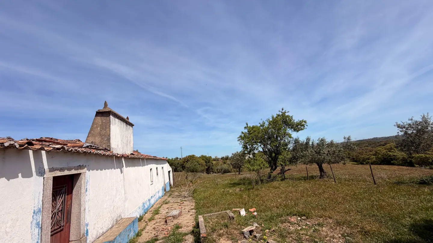Exterior view of a white, one-story house with a red tile roof and a tall chimney, set in a grassy field with trees under a blue sky.