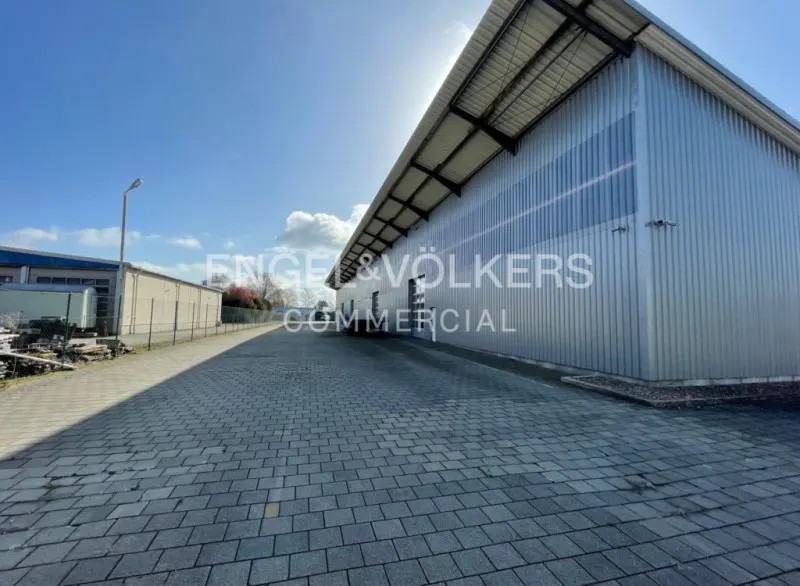 Exterior view of a large, silver metal commercial building with "Engel & Völkers Commercial" logo on the side. Paved driveway in front.