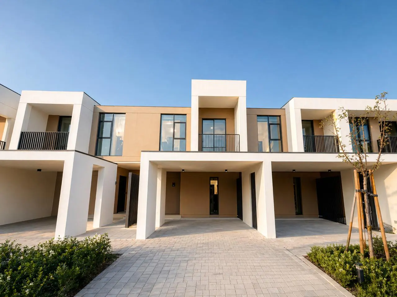 Modern townhouses with beige and white exteriors under a clear blue sky, showcasing a paved driveway and minimalist landscaping.